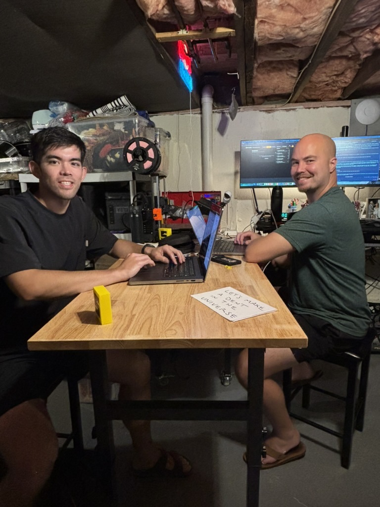 Tim and Daniel at a table in a basement, laptops open, a paper sign between them
